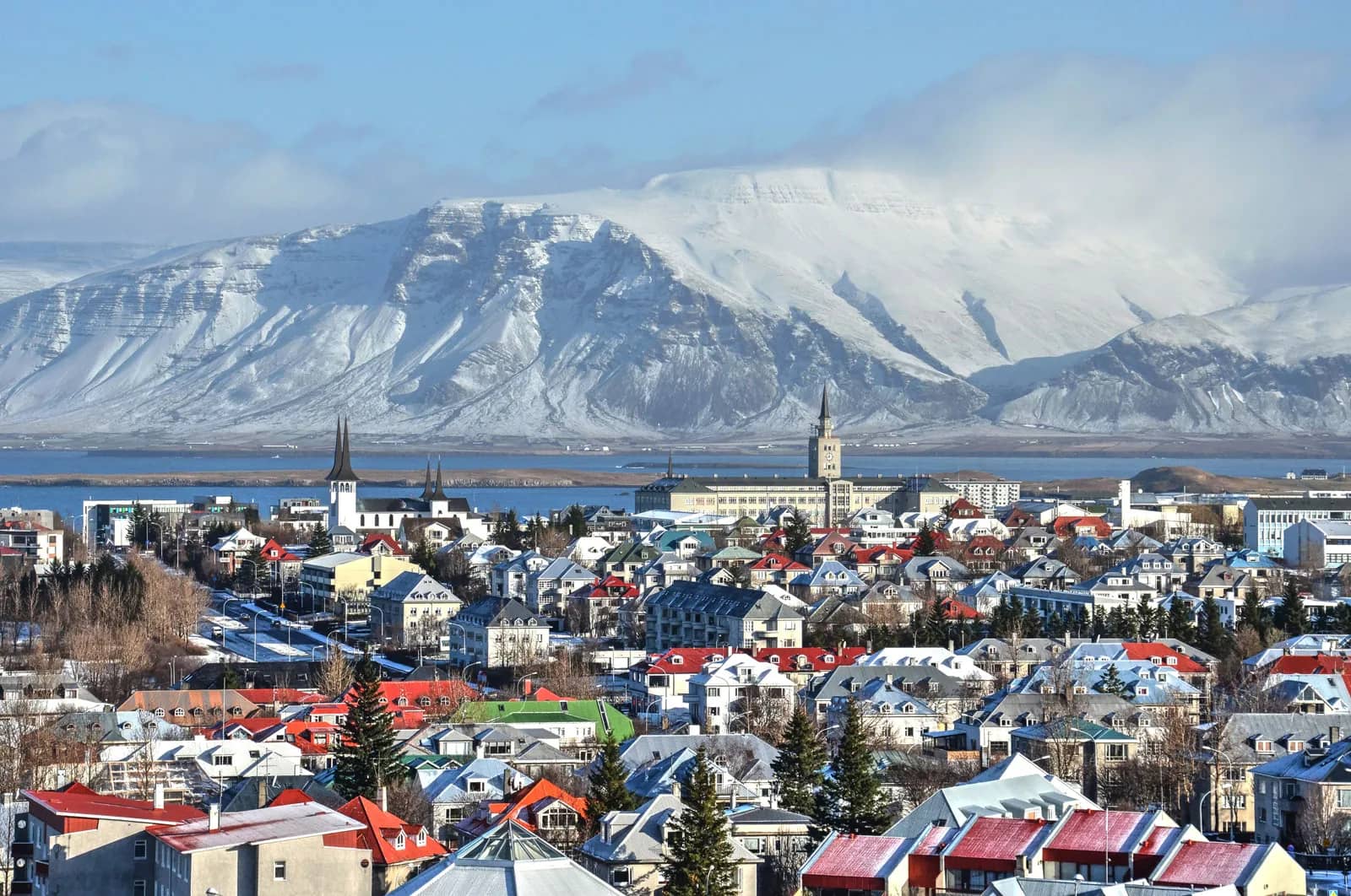 A picturesque view of a small Icelandic town nestled among rolling green hills, with colorful houses dotting the landscape under a clear blue sky. The town is surrounded by natural beauty, including a nearby crystal-clear lake and distant snow-capped mountains, reflecting Iceland's emphasis on clean air and environmental sustainability.