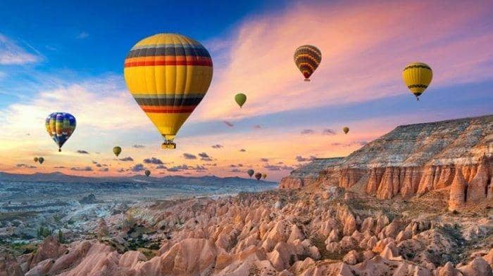 Gambar panorama lembah Cappadocia saat matahari terbit, menampilkan kerucut batu yang ikonik dan balon udara yang melayang di langit yang berwarna-warni, menciptakan pemandangan yang memukau dan unik